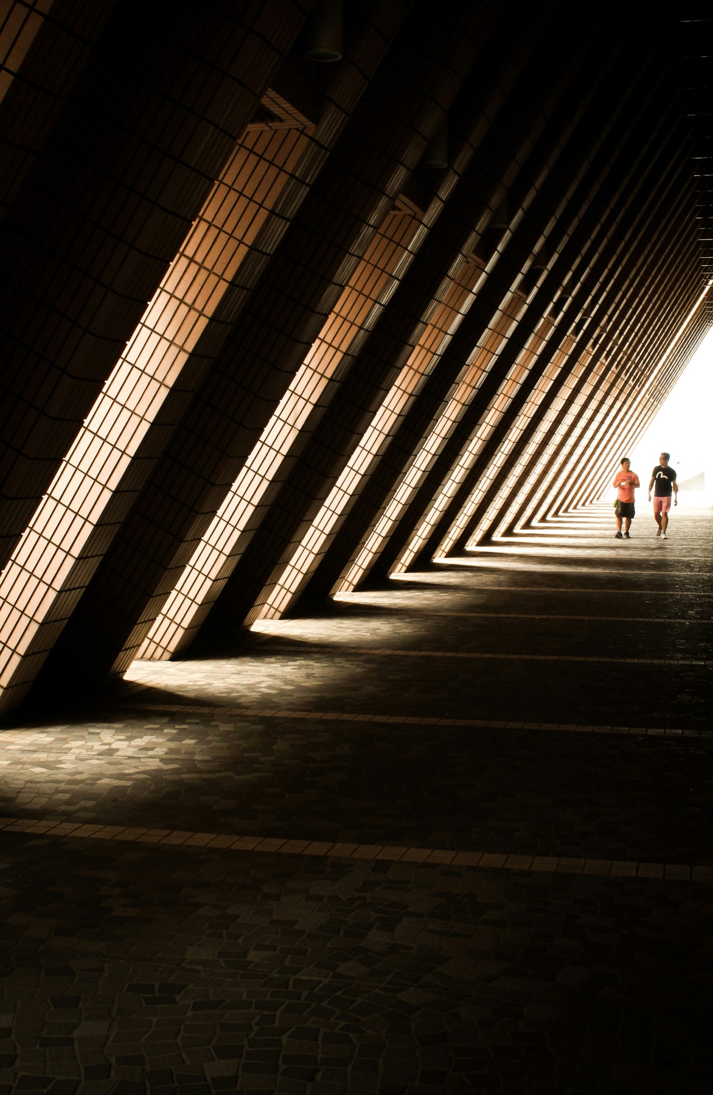 A long colonnade of angled tiled piers, raking light between the columns as two figures walk toward the opening.