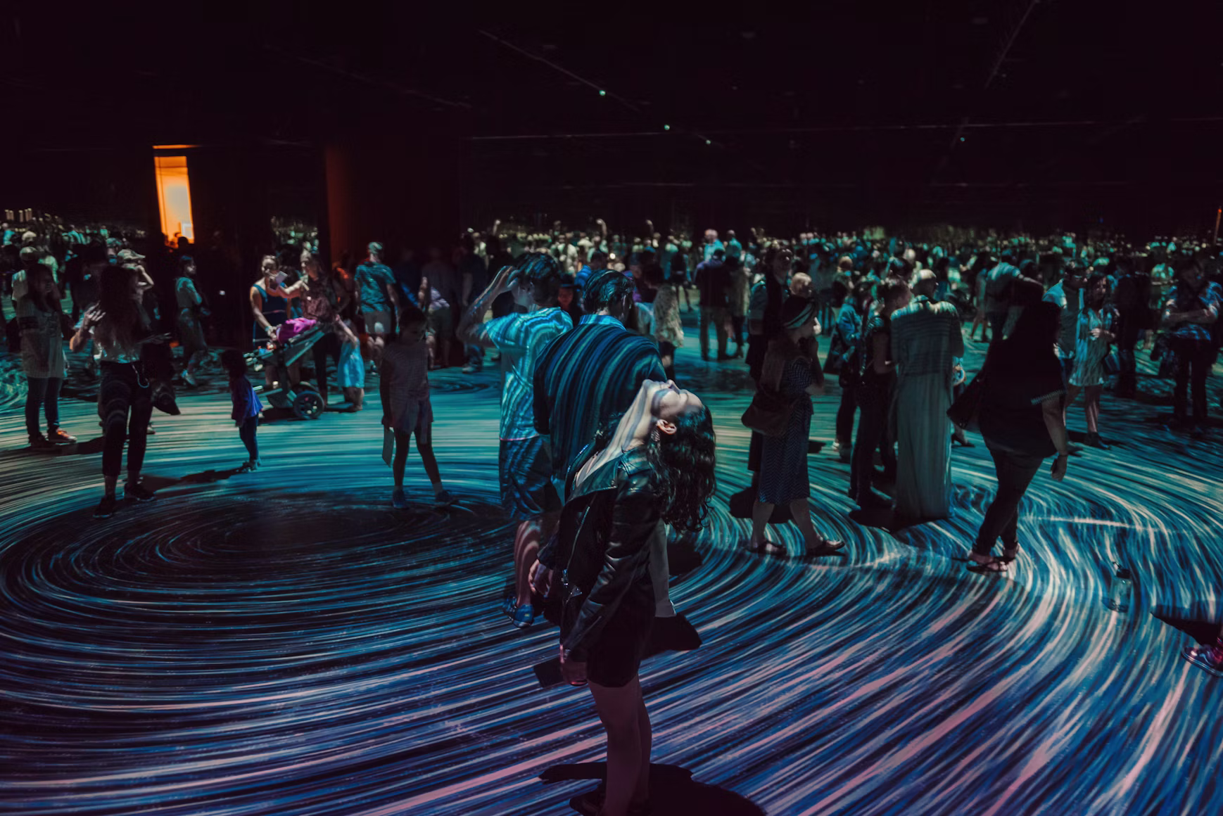Visitors moving through an immersive installation, a vast swirling field of projected light flowing across the floor around them.