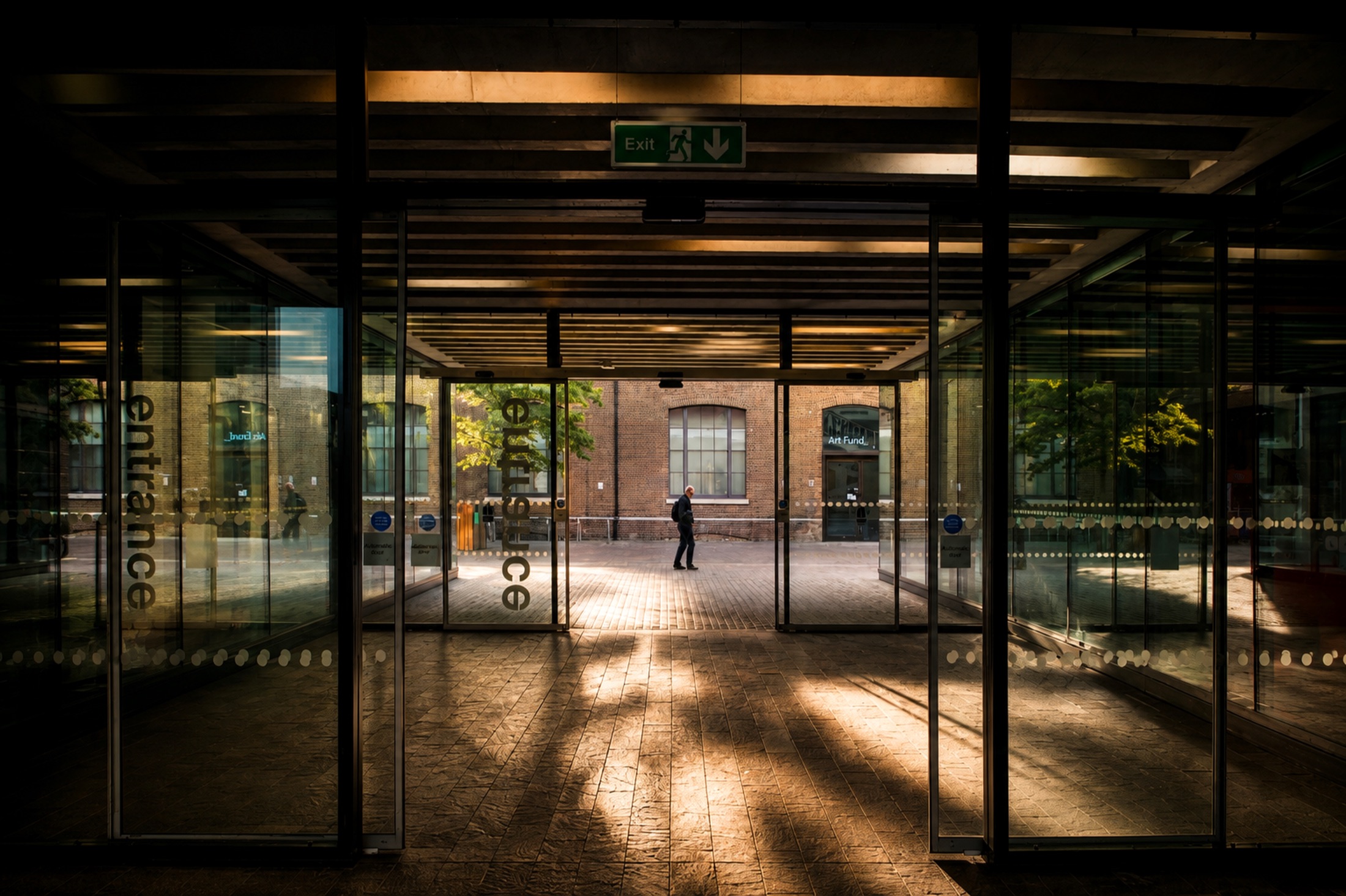 A glazed entrance threshold at dusk, warm light beyond and a single figure approaching.