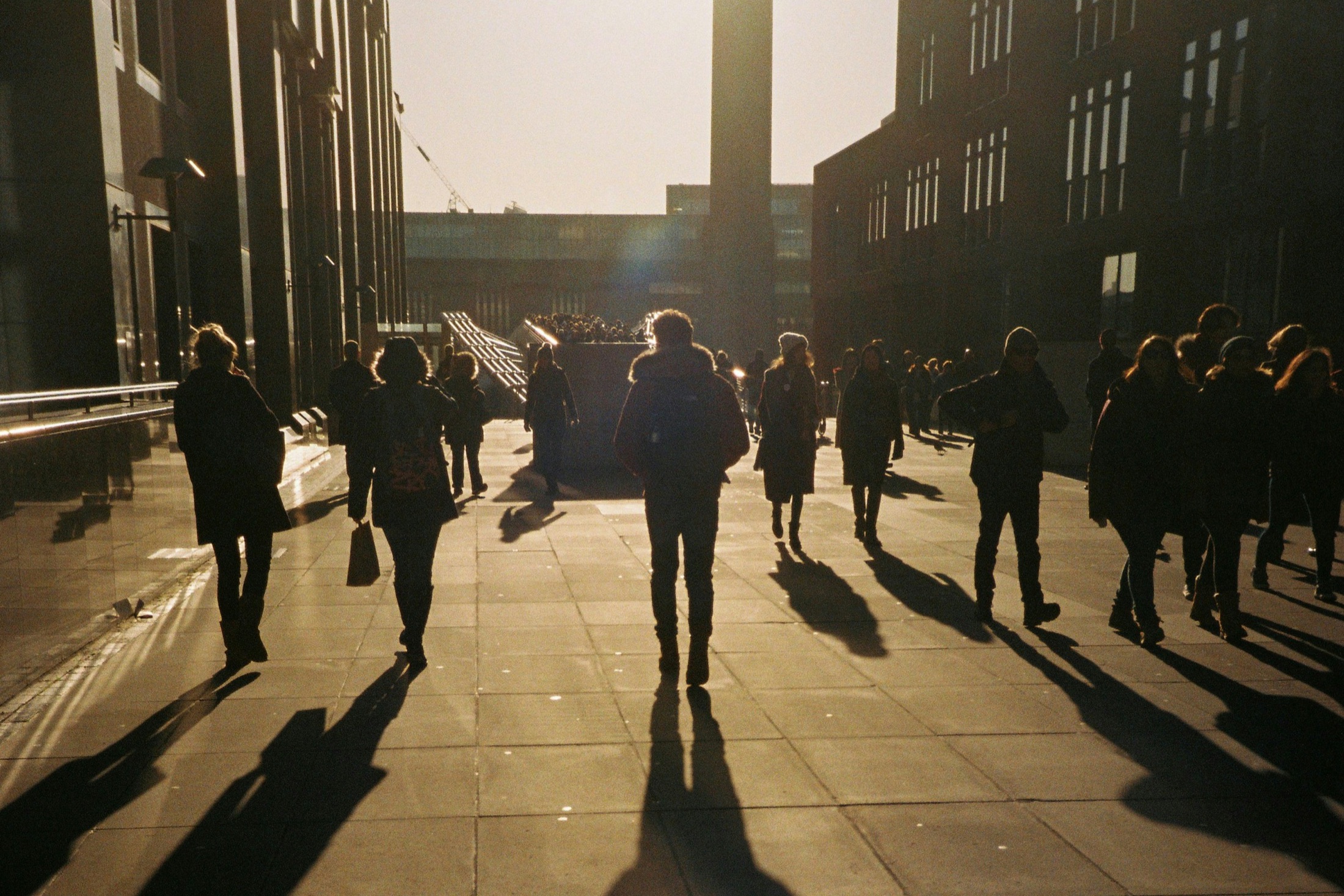 Commuters in silhouette walking through a city plaza in low golden light.
