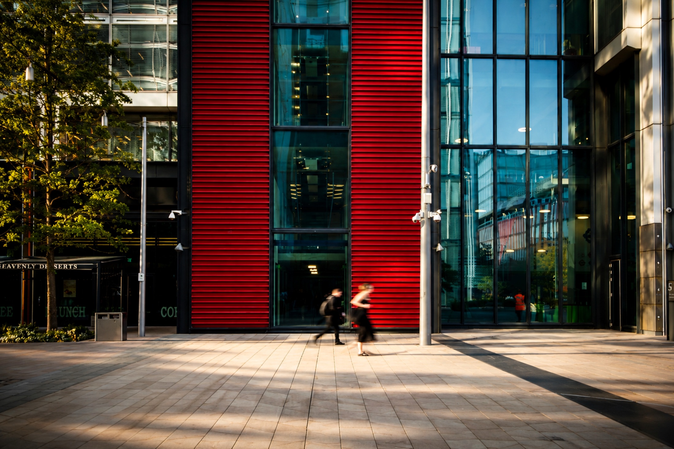 A commercial plaza framed by a red louvred facade, two figures crossing in motion blur.