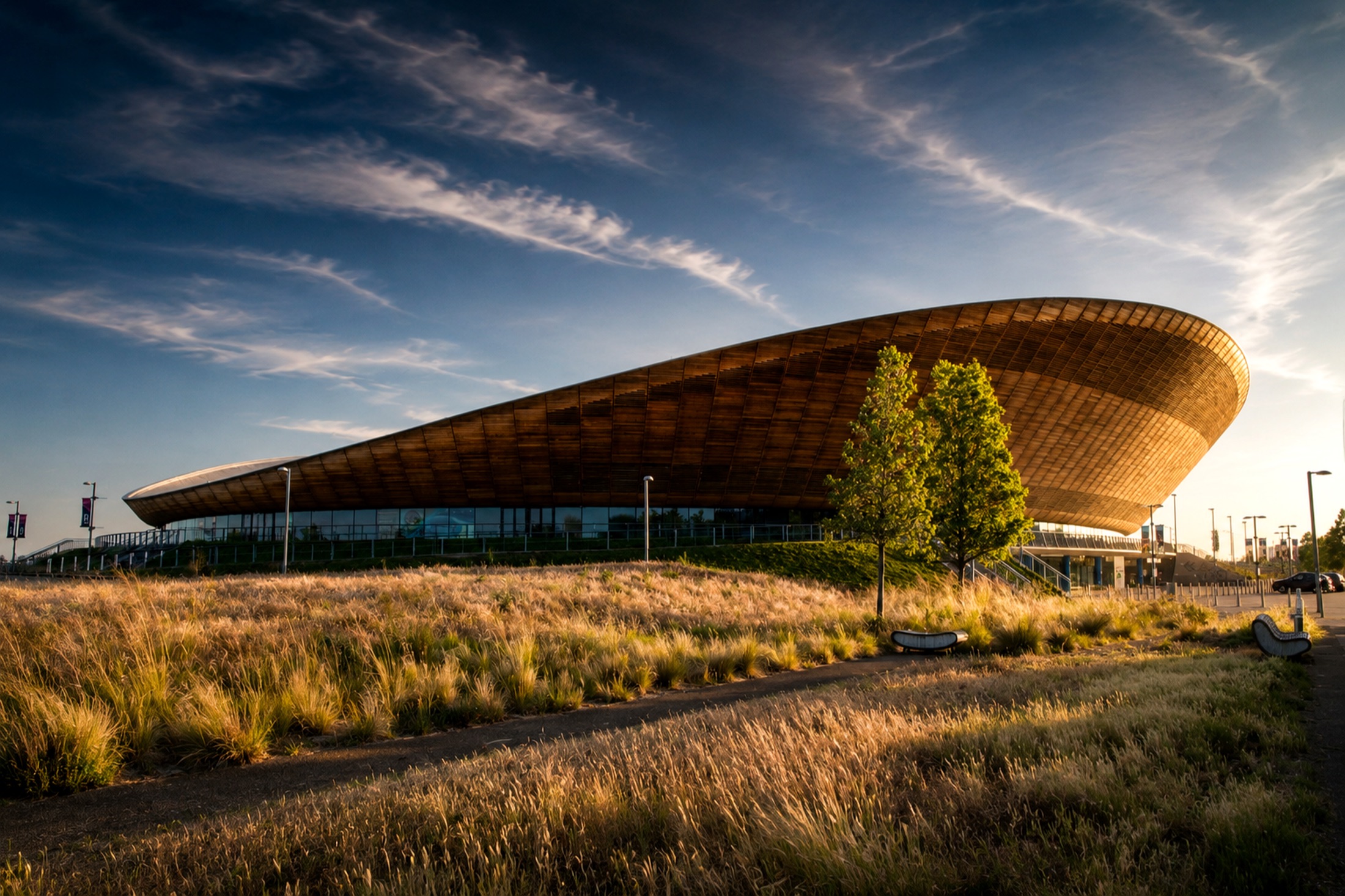 A sweeping timber-roofed velodrome at golden hour, set in long meadow grass.