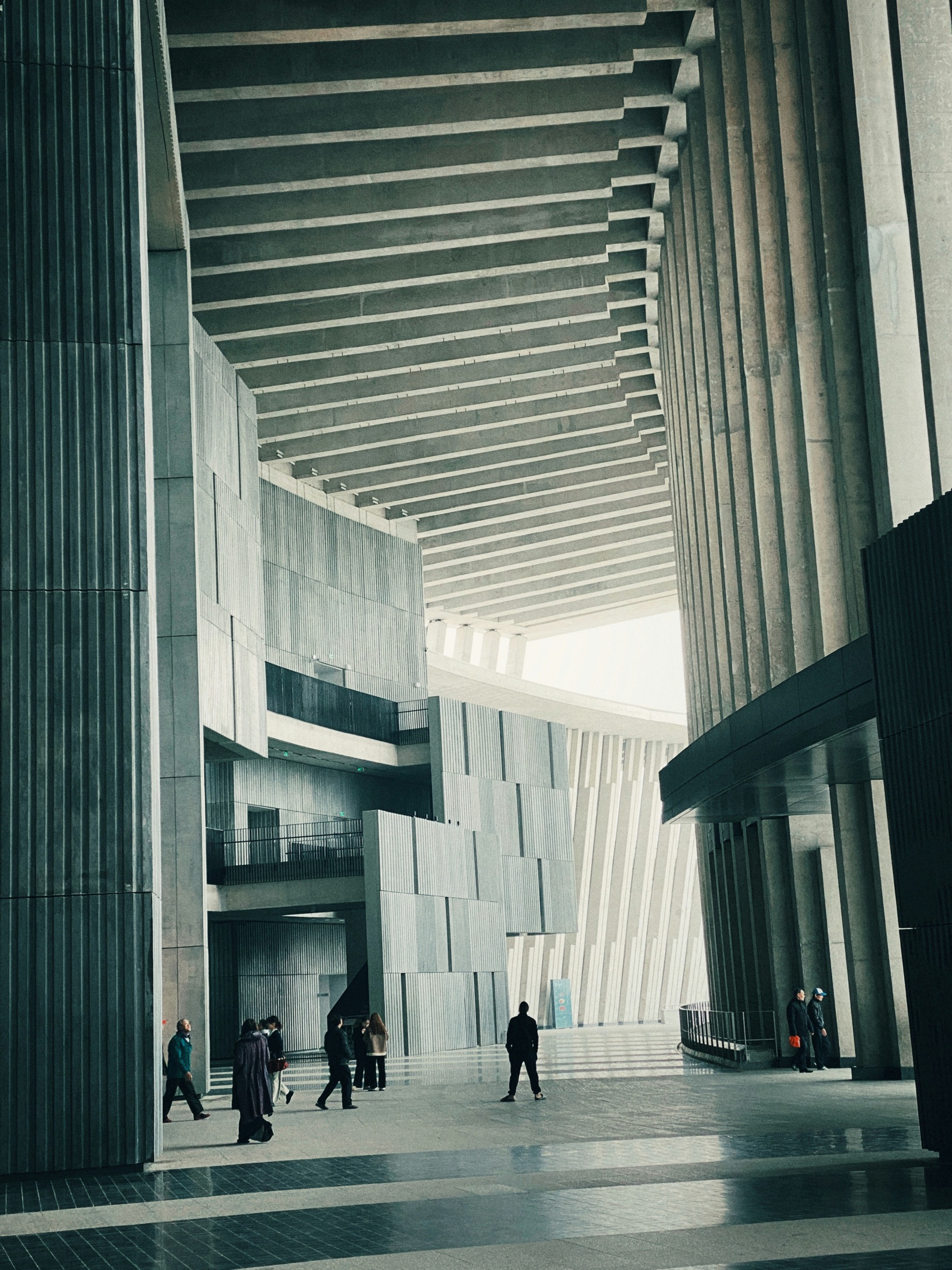 A soaring concrete hall with a slatted ceiling sweeping toward a bright curved opening, tiny figures crossing below.